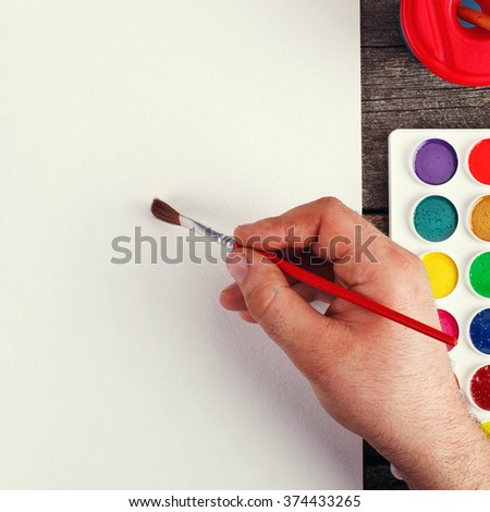 hand with brush painting with watercolor  and blank white paper sheet  on vintage wooden background