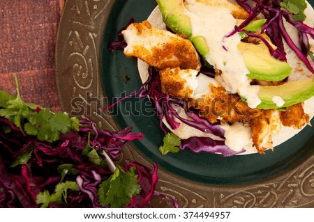 Fresh, home made fish tacos served with avocado and a red cabbage side salad. Presented on a rustic wooden table.
