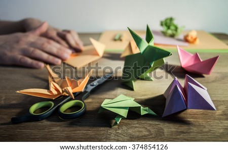 Several origami figures on a wooden table - in the background hands folding colored paper. Royalty-Free Stock Photo #374854126