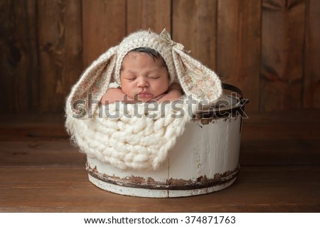 A three week old newborn baby girl sleeping in a little, white wooden bucket. She is wearing a cream colored bunny rabbit bonnet. Shot in the studio on a wood background.
