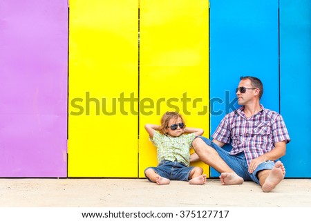 Father and son relaxing near the house at the day time. They sitting near are the colorful wall. Concept of friendly family.
