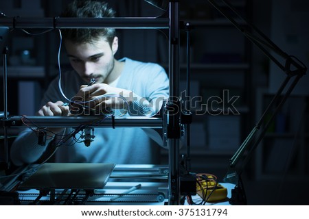 Young engineer working at night in the lab, he is adjusting a 3D printer's components, technology and engineering concept Royalty-Free Stock Photo #375131794