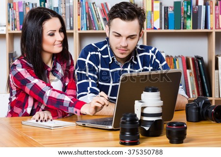 Attractive boy and beautiful brunette girl wearing in plaid shirts sitting with laptop, notebook, photo camera and object glasses, books at background.