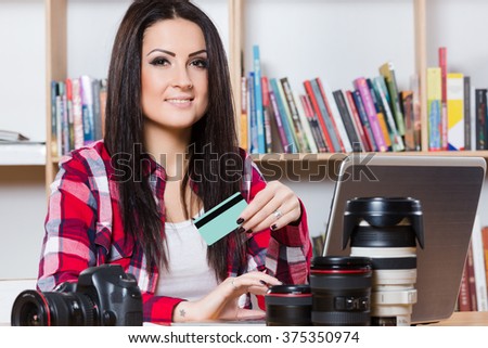 Beautiful young brunette wearing red plaid shirt sitting with laptop, photo camera and object glasses, holding credit card, a lot of books at background.