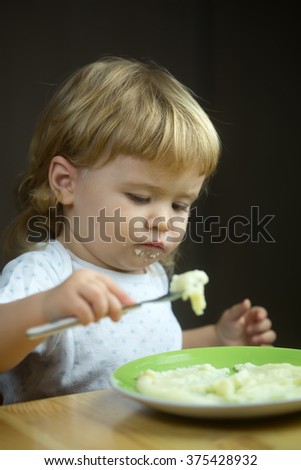 Closeup portrait view of one adorable cute small baby boy with blonde hair eating healthy food of porridge or coocked semolina from plate with spoon in hand indoor, vertical picture