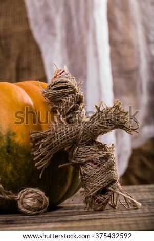 Closeup photo of scary cross shaped Halloween witching toy with clew of twine near orange pumpkin with green blotch on wooden table on white and brown cloth background, vertical picture