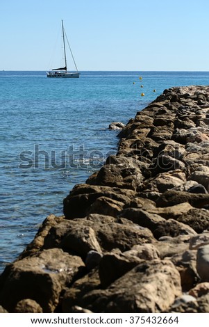 Photo closeup of stone pier coast and one classic yacht sailing boat vessel offshore in calm blue sea silhouetted against clear sky day time on beautiful seascape background, vertical picture