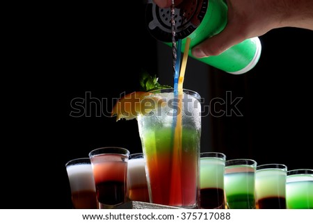Beautiful delicious sweet bright red and green alcohol cocktail in transparent drinking glass with sipping straws pouring from shaker near many colorful shots on black background closeup, horizontal 