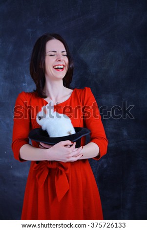 Teacher of of Magic. Young beautiful girl in a red dress holds a black illusionist hat with a white rabbit inside it with a blackboard background.