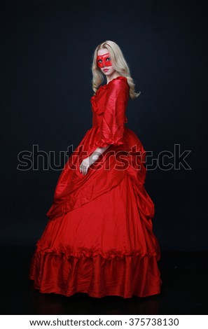 full length portrait of a beautiful blonde woman wearing a red lace mask and a historical red silk, victorian era ball gown.
