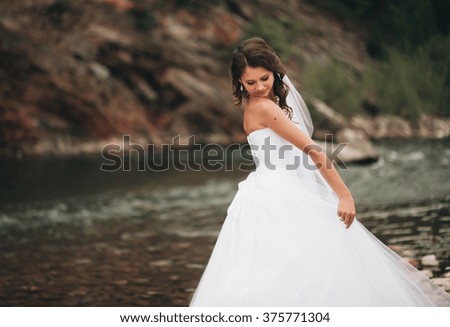 Beautiful luxury young bride in long white wedding dress and veil standing near river with mountains on background