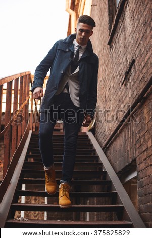 Young hipster man on stairs. A man wearing in a blue jacket , a white shirt, blue jeans and yellow boots Royalty-Free Stock Photo #375825829