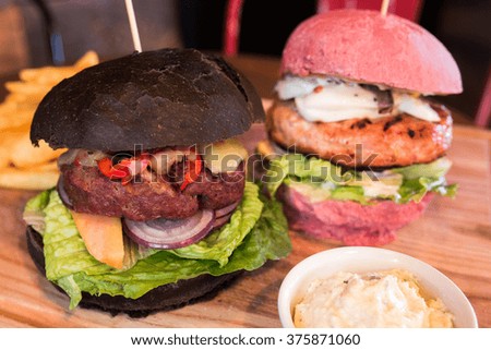 Two burgers. One with turkey, lettuce, avocado, egg, anchovies and caesar dressing in pink bun and second one with beef, red bean paste, chili pepper, lettuce, papaya and cheddar cheese in black bun.
