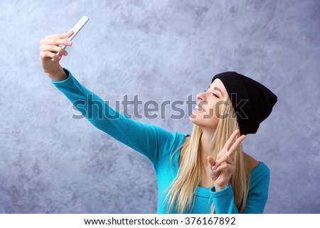 Young girl in black hat taking photo of her self with smart phone on grey wall background