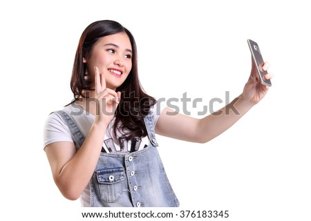 Cheerful Asian girl taking selfie with her smartphone camera and making victory hand sign, isolated on white background