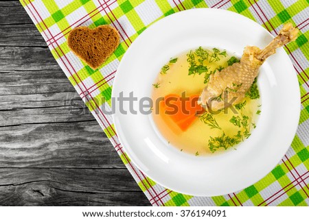 Fresh clear soup with carrot and chicken leg decorated with dill in a wide rim round plate on a napkin  on an old wooden table, top view