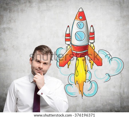 A young man with a hand to the chin standing in front of a concrete wall with a coloured image of a rocket to the right. Concept of launching a new project.