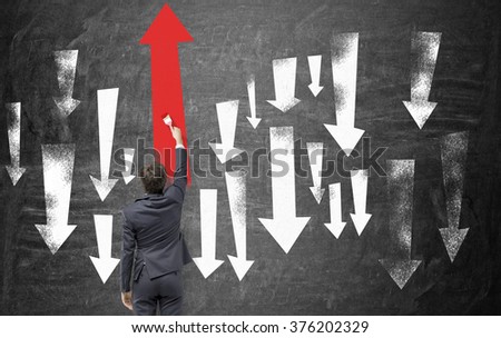 A businessman painting big up and down arrows on the blackboard, the biggest of them is a red one up. Back view. Concept of moving up.