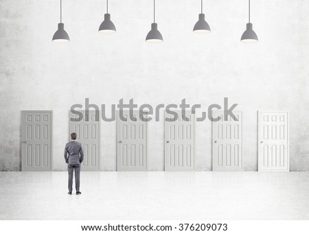 A young businessman with hands in pockets standing in a room with numerous closed doors. Five lamps above. Back view. Concrete background. Concept of finding a way out.