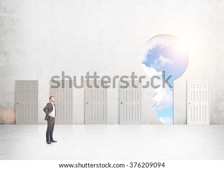 A young businessman with hands on hips standing in a room with numerous closed doors, a keyhole with sky seen instead of one. Side view. Concrete background. Concept of finding a way out.