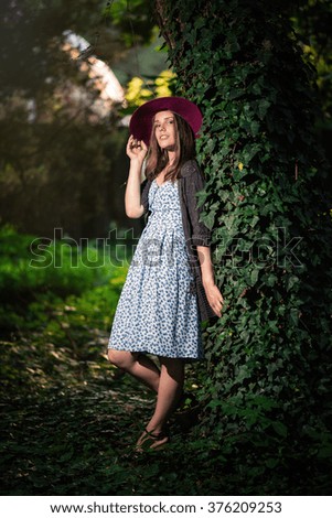 pretty slim girl with long hair in an elegant hat in a simple flowered dress in warm jacket with a camera in hand walking in the summer in the park at sunset