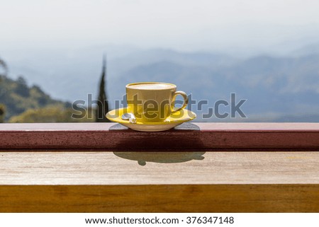 This image focus on a cup of coffee on the outdoor banister and have mountain view blurry at far away.