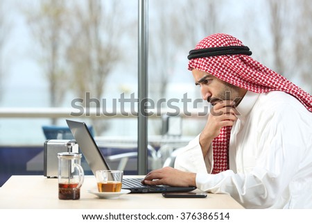 Side view of an arab saudi man worried working with a laptop in a coffee shop interior with the terrace in the background