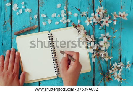 woman writing on blank notebook next to spring white cherry blossoms tree on vintage wooden table. 