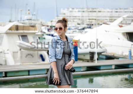 Picture of beautiful young lady holding camera standing in harbour. Portrait of elegant photographer shooting yachts on summer sunny outdoor background.