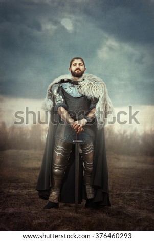 The courageous knight dressed in chain armor standing in a field leaning on his sword. Royalty-Free Stock Photo #376460293