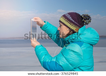 Smiling woman uses a tablet pc, making photo, during a walk along the coast in sunny winter day. Toned image.