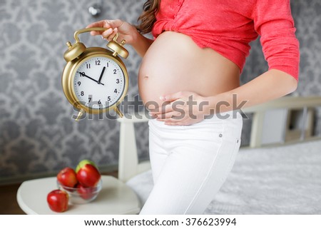 Pregnant woman holding alarm clock. Pregnant woman's belly. closeup of a pregnant woman holding an alarm-clock near her belly at home in bedroom. 