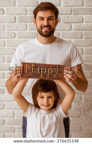 Portrait of a handsome young father and his cute little son holding a wooden plate "happy", looking in camera and smiling. Both in white t-shirts and jeans, standing against white brick wall.