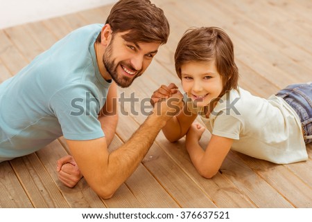 Top view of handsome young father in casual clothes and his cute little son competing in arm wrestling while lying on a wooden floor in the room