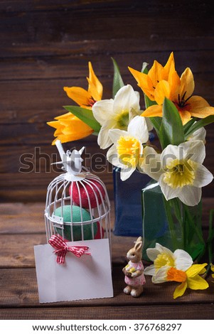 Colorful easter eggs in bird cage, fresh tulips and narcissus flowers  in vases, empty tag  on aged dark wooden background. Easter background. Place for text.