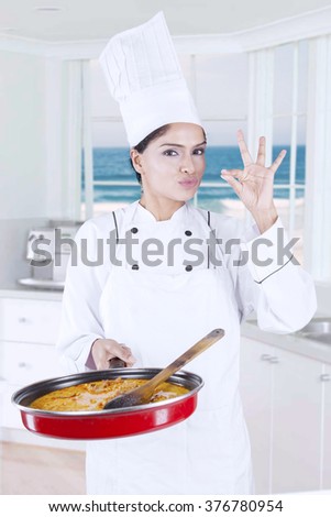 Portrait of indian female chef showing ok sign while holding pan and standing in the kitchen