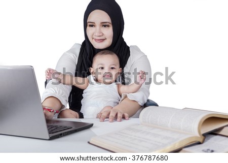 Picture of multi-tasking mother using laptop computer with her baby and books on desk, isolated on white background