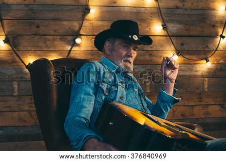 Smoking senior country and western guitarist with beard sitting in chair.