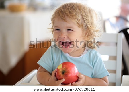 Laughing cute fair-haired blond hazel-eyed kid little child baby boy sitting in highchair and eating big red apple fruit portrait on blurred background, horizontal picture