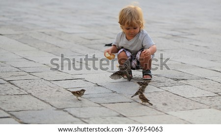Cute fair-haired blond kid tiny little child baby boy feeding sparrows with bun sitting on haunches on flag-stone pavement cityscape on blurred grey background, horizontal picture