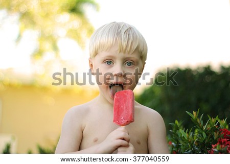 A young boy child is outside by palm trees on a summer day eating a red strawberry fruit popsicle for a treat.