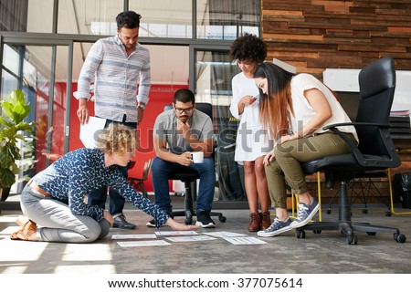 Creative people looking at project plan laid out on floor. Mixed race business associates discussing new project plan in modern office. Royalty-Free Stock Photo #377075614