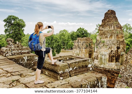 Young female tourist with blue backpack taking picture with smartphone of the gopura from the top of ancient Pre Rup temple Khmer in Angkor on blue sky background. Siem Reap, Cambodia.