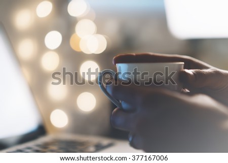 close-up of a cup of coffee or tea in women hands on a light background bokeh and flare with an open laptop