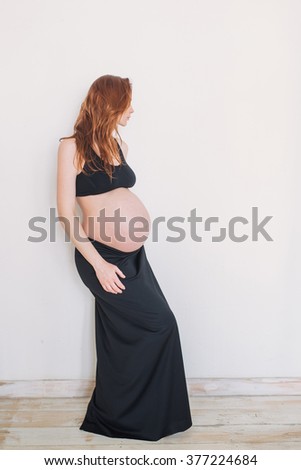 Pregnant woman with red hair in black clothes on a white wall