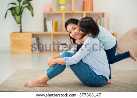 Happy young mother sitting on the floor when her daughter kissing her from behind