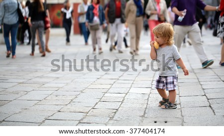 Photo closeup of cute fair-haired blond kid tiny little child baby boy eating bun standing on flag-stone pavement in crowd cityscape on blurred grey background, horizontal picture