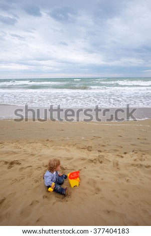Kid cute fair-haired blond tiny little child baby boy sitting on sand and playing with yellow toy trowel bucket on beach on windy murky day on blurred seascape background, vertical picture