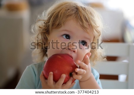 Cute fair-haired blond hazel-eyed kid little child baby boy biting and eating big red apple fruit portrait on blurred background, horizontal picture