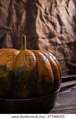 One raw flat round orange squash with smooth segmented surface green blotch and stem on pan with black handle on wooden table on burlap background, vertical picture